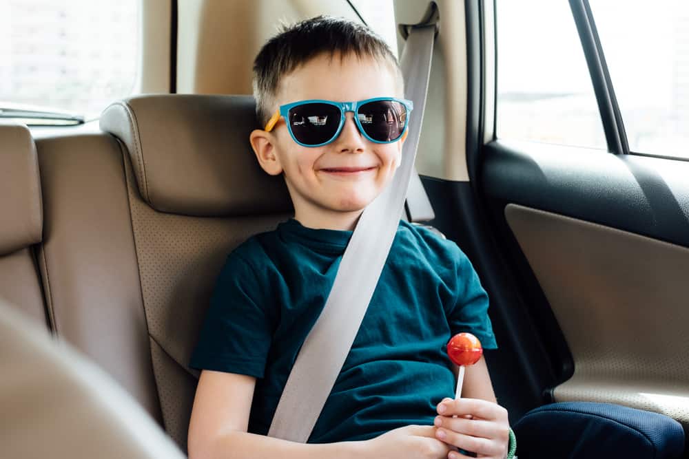 happy child with sunglasses and lollipop inside car wearing his seat belt