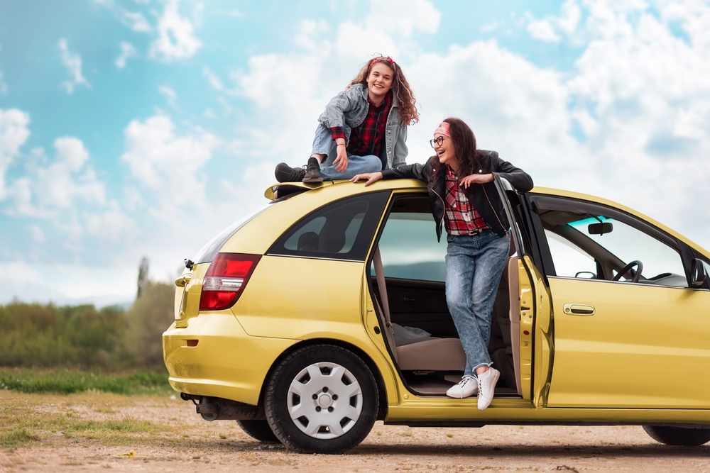 Two friends on a yellow hatchback, one sitting on the roof and the other leaning out the open door, celebrating a sunny getaway drive—Washington Road Trip Tips.