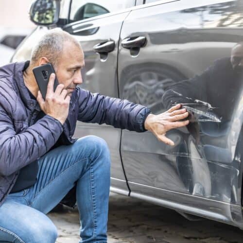 A man examines damage to his car while on the phone to his car insurance agency