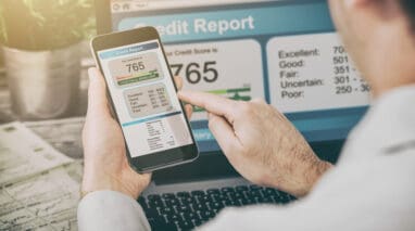 man looks at his credit score on his phone with laptop in background