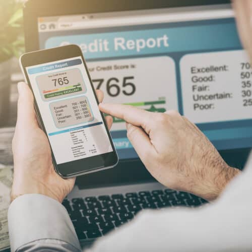 man looks at his credit score on his phone with laptop in background