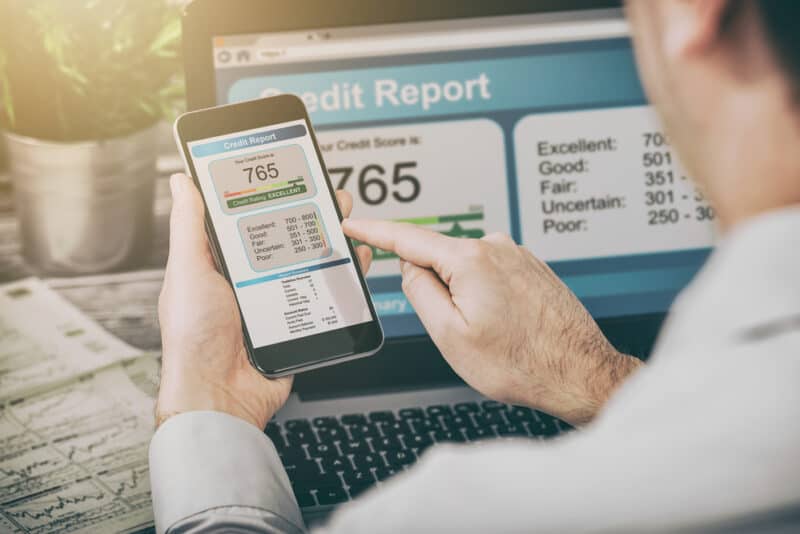 man looks at his credit score on his phone with laptop in background