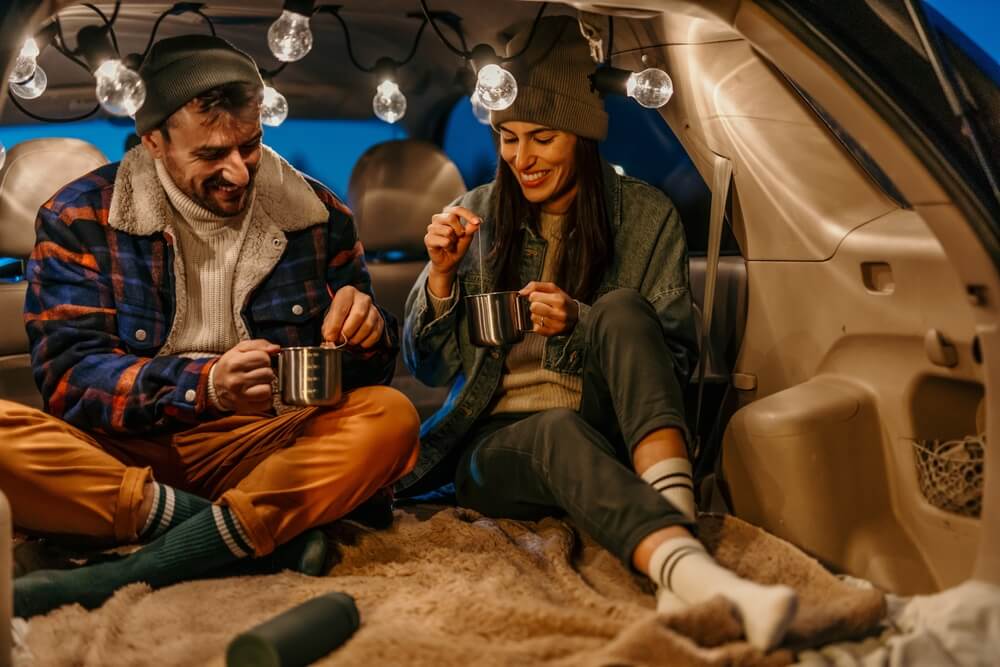 Two people sharing hot drinks from the back of a vehicle, taking in a scenic car camping getaway.