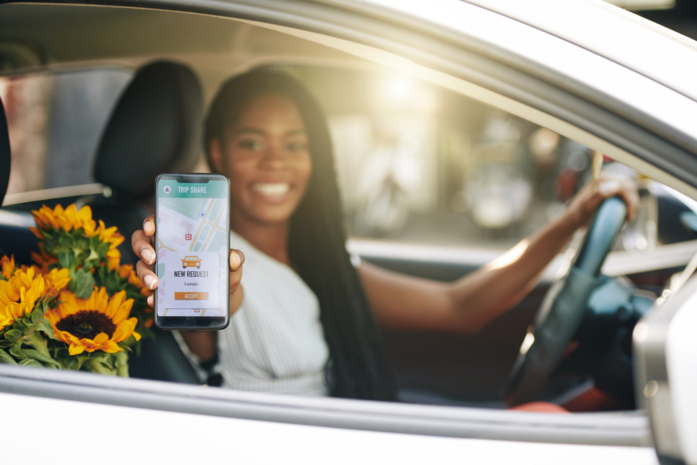 Young woman, smiling, behind the wheel, holds phone towards camera to show a ride request has come in - cheap rideshare insurance in Washington.