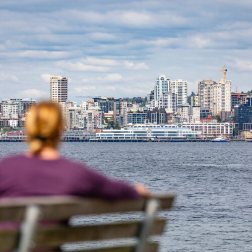 Woman sits on bench gazing across water at Seattle Space Needle - cheap car insurance in Washington.