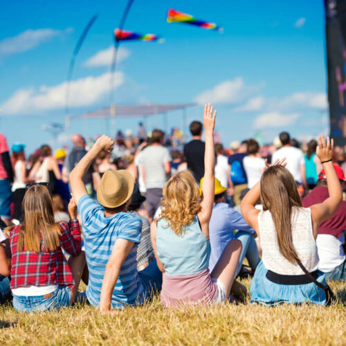 Large group of people sitting on grass watching and listening to music festival.