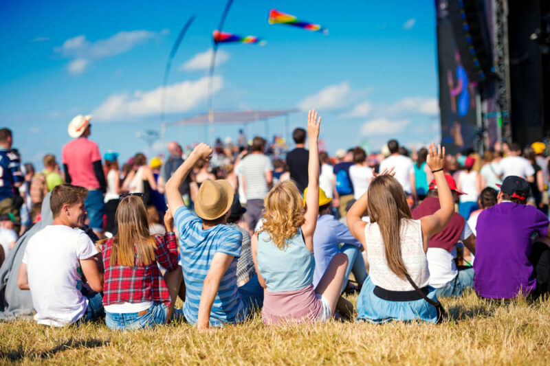 Large group of people sitting on grass watching and listening to music festival.