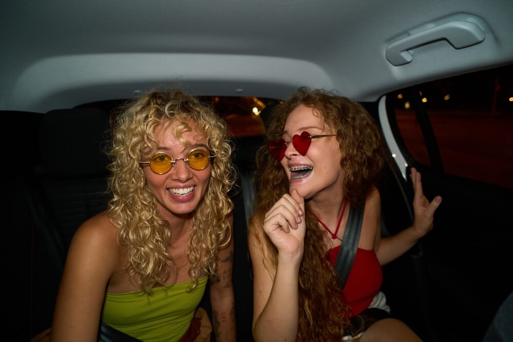 Two women laugh in the back seat of a car at night, wearing colorful sunglasses and seat belts, capturing a fun moment while thinking about how to insure someone else's car.