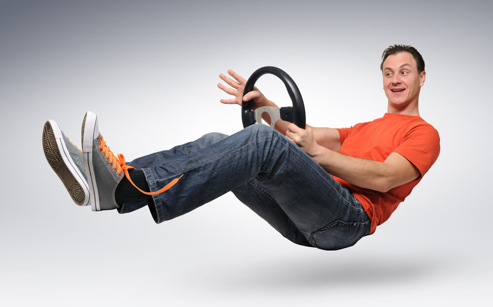 Man playfully “floating” while gripping a steering wheel against a plain background, illustrating how to insure someone else’s car without the usual setup.