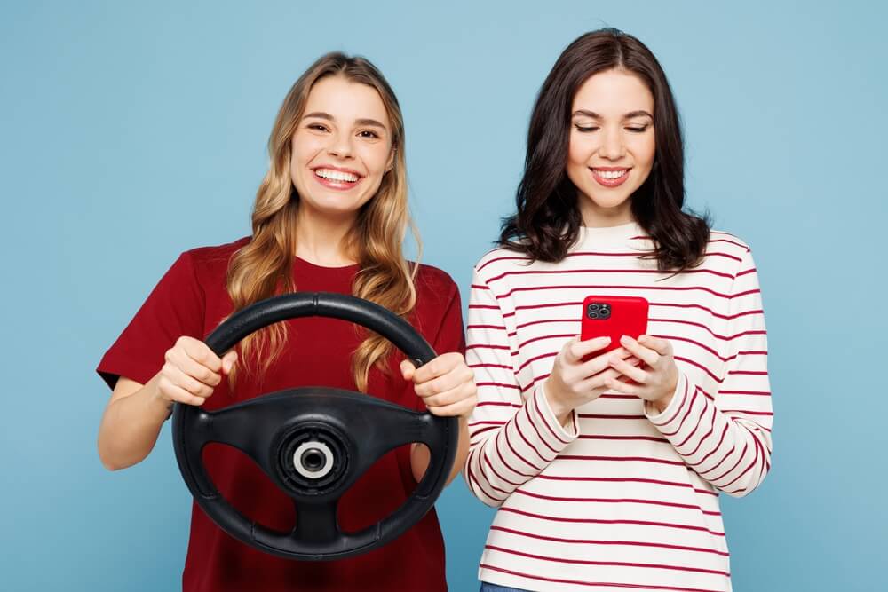 Two women stand against a blue background, one smiling while holding a steering wheel and the other checking her phone, illustrating how to Insure Someone Else's Car.