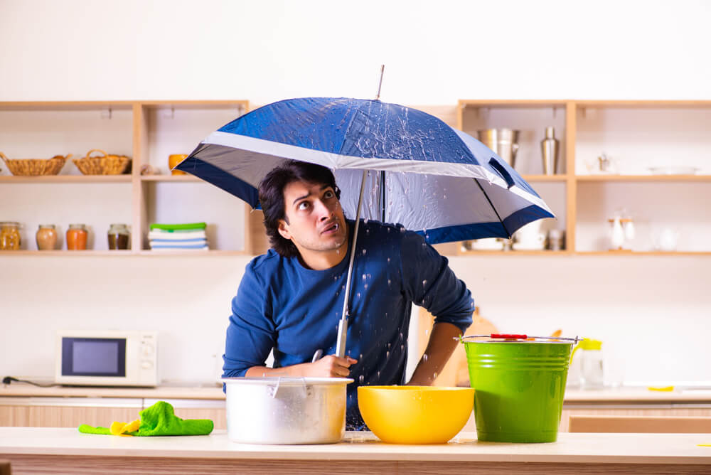 Man standing in kitchen with umbrella and buckets for leaking roof - cheap home insurance in Washington.
