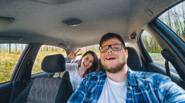 Man laughing behind wheel of car while woman looks on from back seat, concept of insuring someone else's car - cheap car insurance in Washington.