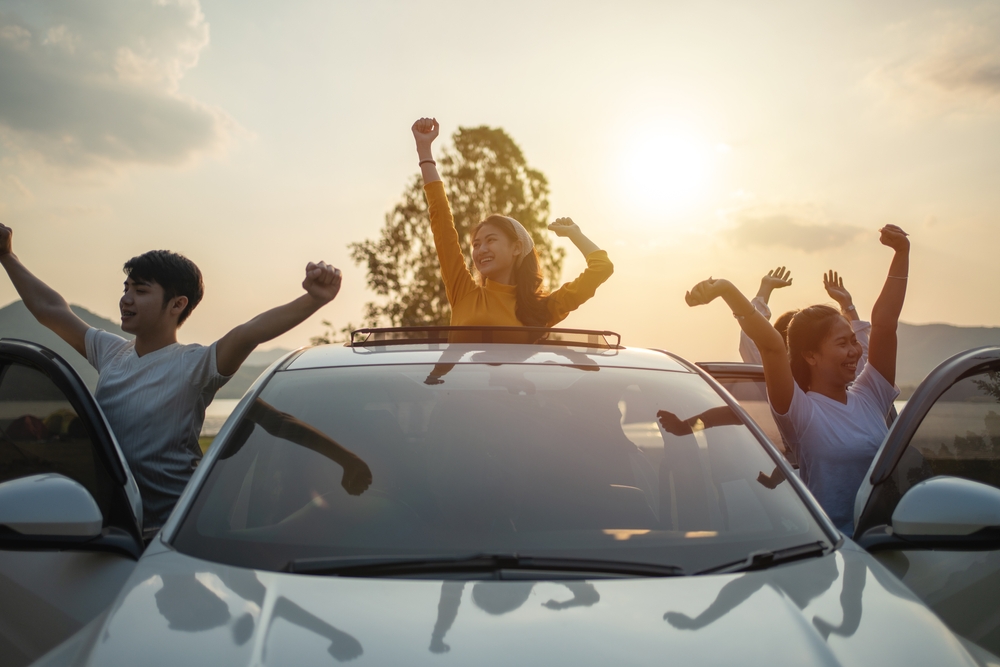 Friends celebrating around a car during a road trip at sunset.