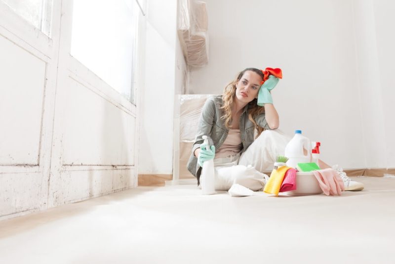 Tired and disgusted woman, cleaning mold from home in Washington State, using house cleaning products and wearing gloves for remediating and removing mold before painting, does home insurance cover mold?