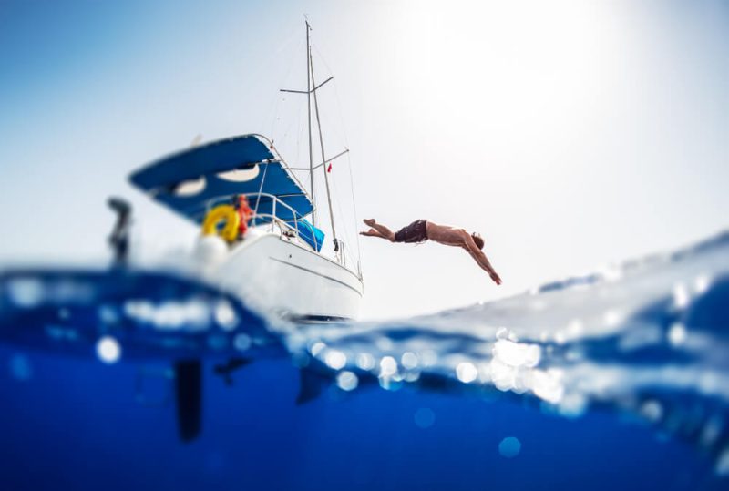 Young man jumps into the sea from the sail boat, image split over under image with the crystal clear sea, cheap boat insurance for protection.