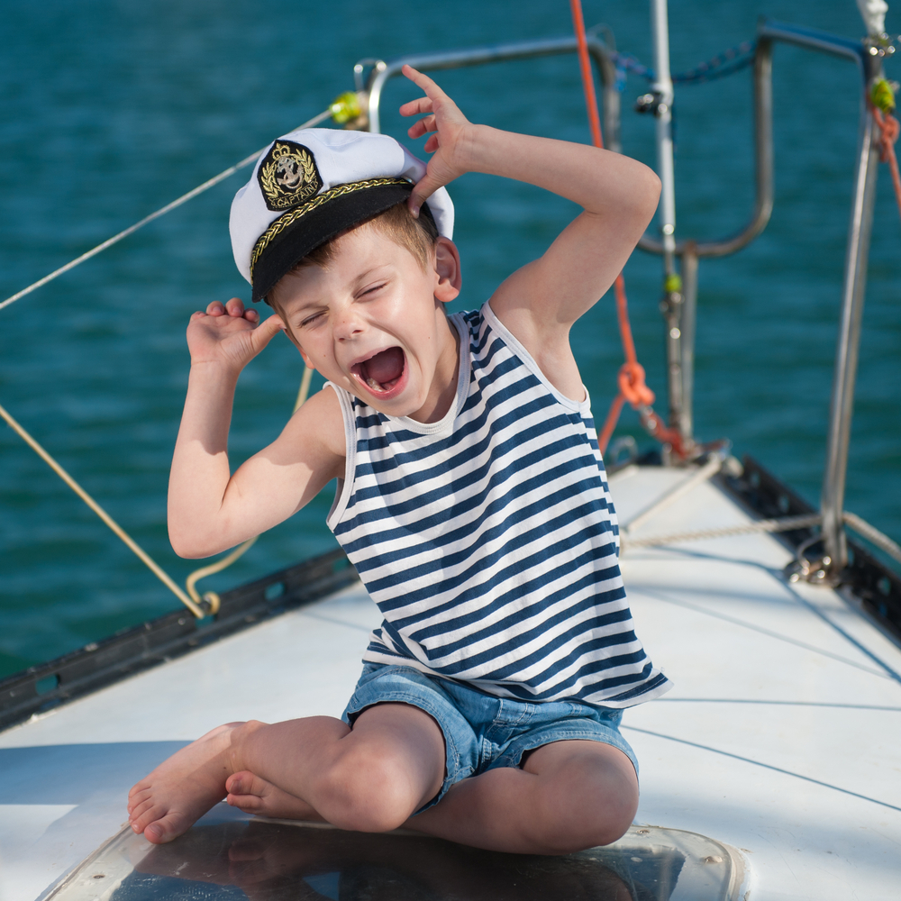 Funny little boy wearing captain hat sitting aboard boat with water behind in summer, cheap boat insurance in Washington for protection.