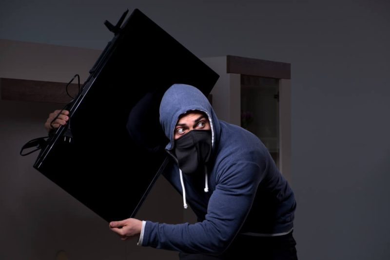 Burglar in black balaclava stealing tv set from house. Burglar or intruder stealing in property apartment. Need for cheap renters insurance in Washington State.