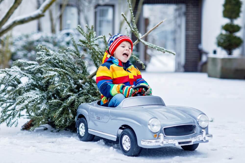 A boy smiling and playing on a toy car in the middle of the snow with a pine tree behind him — a reminder of why winter auto insurance tips are important for safe and worry-free driving.