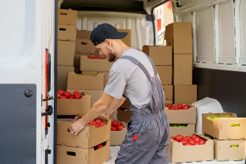 Harvest delivery to fruit market on food truck as male worker carrying boxes of vegetables. Transport service to grocery trade from farm factory. 