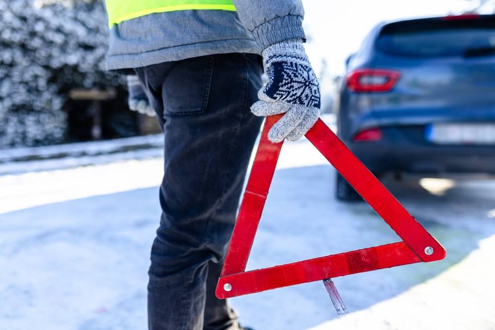 A close-up of a man's hand holding a red warning triangle on a snowy road underscores how winter breakdowns make commercial auto insurance essential.
