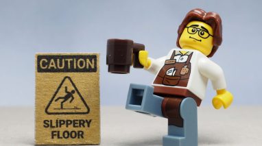 Mini toy of barista man serving in a cafeteria, holding a cup of coffee, about to slip on a wet floor next to a wet floor sign, highlighting business insurance in Washington.
