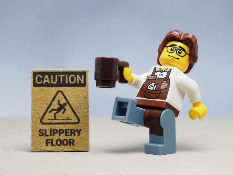 Mini toy of barista man serving in a cafeteria, holding a cup of coffee, about to slip on a wet floor next to a wet floor sign, highlighting business insurance in Washington.