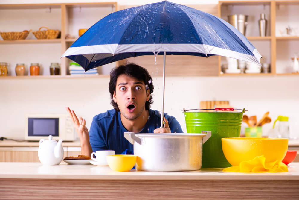 Shocked man holds an umbrella indoors as water drips from the ceiling into buckets and pots on a kitchen counter, highlighting common insurance gaps.