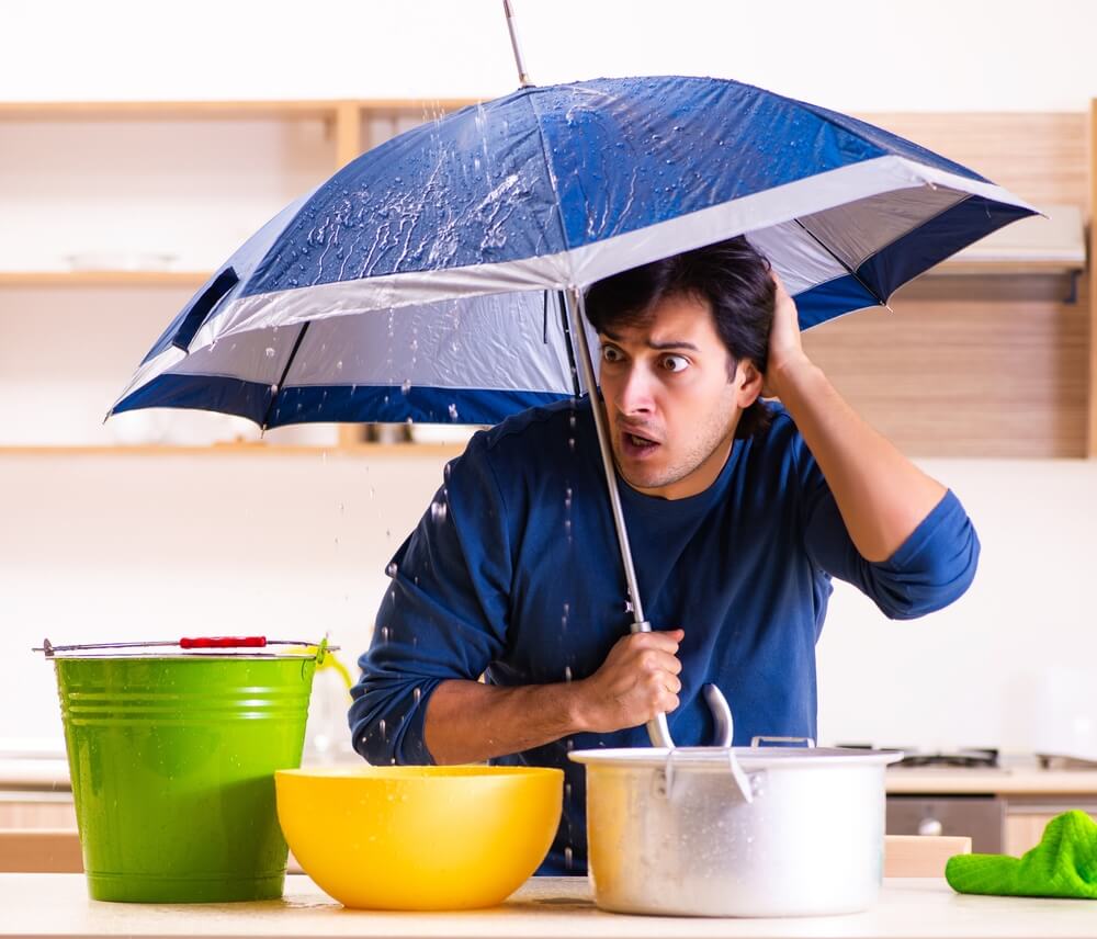 Man looks shocked while holding an umbrella indoors under a ceiling leak, with buckets and pots catching dripping water, reminding renters to review insurance policy after water damage.