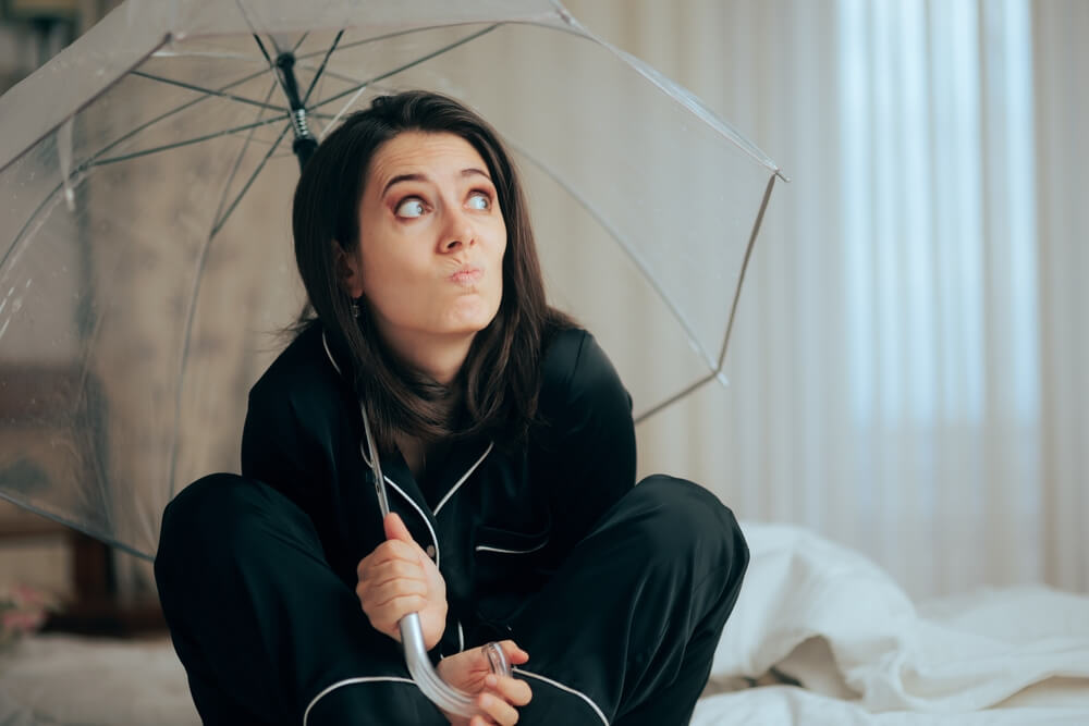 Woman in dark pajamas sits on a bed holding a clear umbrella indoors and looks up with a worried expression, questioning home insurance myths about what water damage is actually covered.