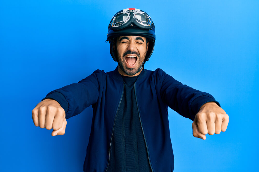 Smiling man wearing a motorcycle helmet and goggles pretends to ride with his hands outstretched against a blue studio background, highlighting Motorcycle Insurance in Washington.