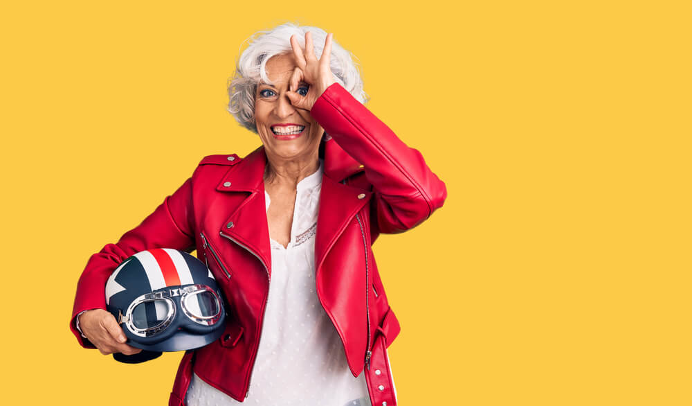 Smiling older woman in a red jacket holds a motorcycle helmet on a yellow background, a playful nod to motorcycle insurance in washington.