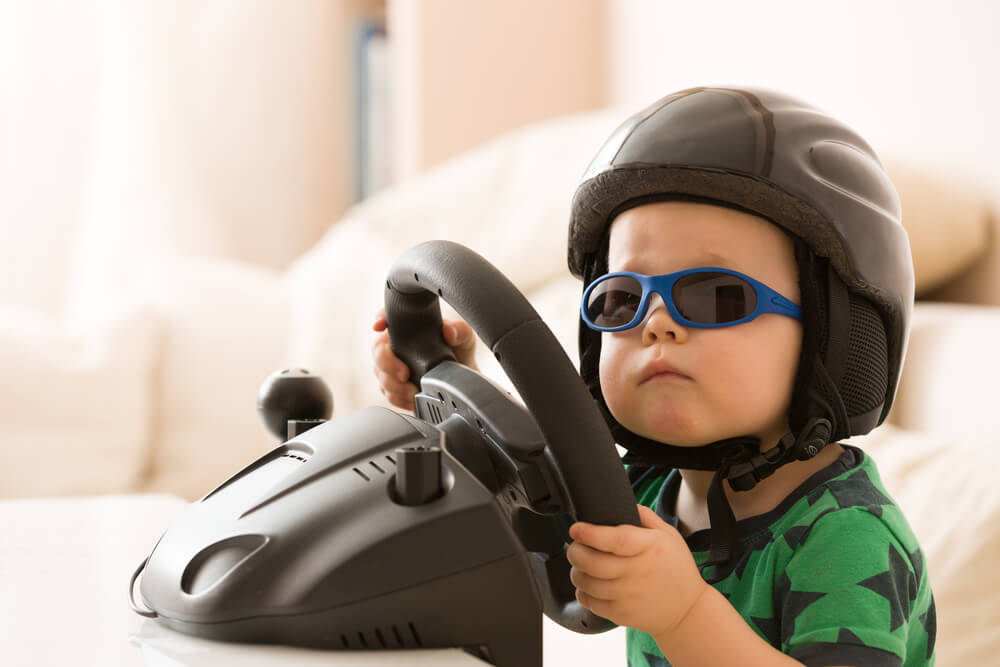 Child wearing a helmet and sunglasses plays with a driving simulator wheel, introducing car technology in a fun way.