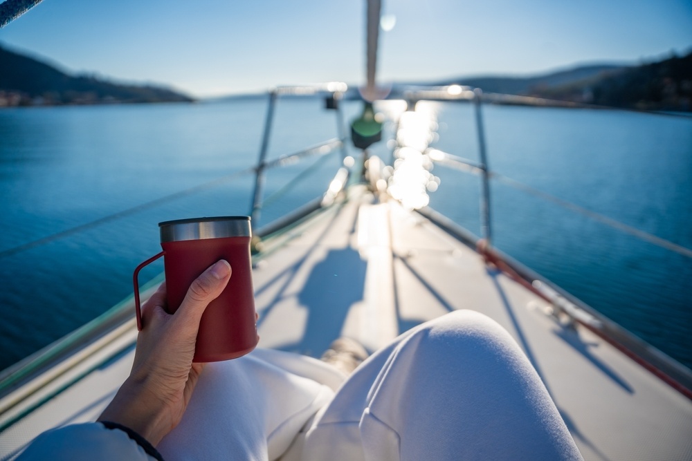 Person holding a red travel mug while relaxing aboard a sailboat on calm blue water during a sunny day.